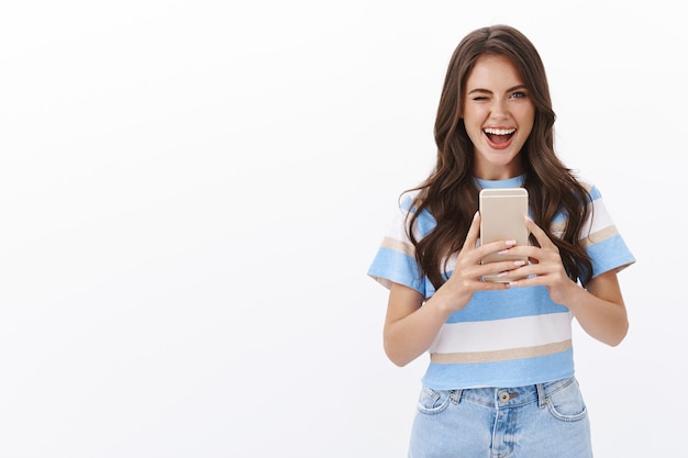 Mujer moderna con estilo descarado que toma selfie en el espejo, guiñando un ojo ambicioso y coqueto mostrar la lengua, sonriendo encantada, prueba la nueva cámara del teléfono inteligente, fotografiando cerca de la pared blanca