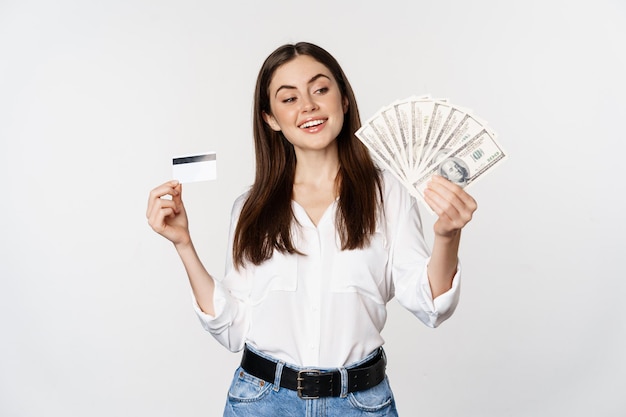 Foto gratuita mujer moderna feliz con dinero y tarjeta de crédito en las manos, sonriendo complacida, de pie sobre fondo blanco. copia espacio