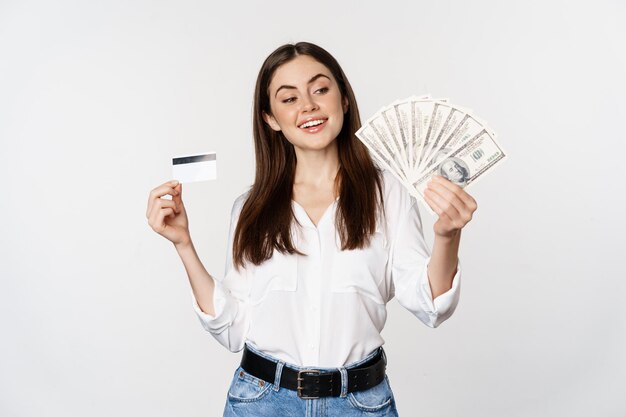 Mujer moderna feliz con dinero y tarjeta de crédito en las manos, sonriendo complacida, de pie sobre fondo blanco. copia espacio