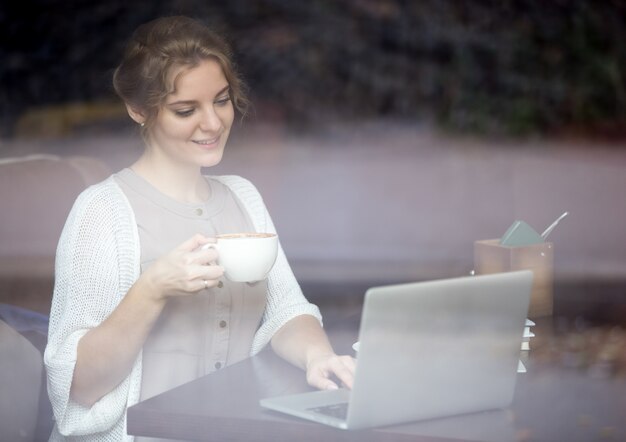 Mujer moderna que trabaja en la computadora portátil en cafetería. Disparo a través de la ventana