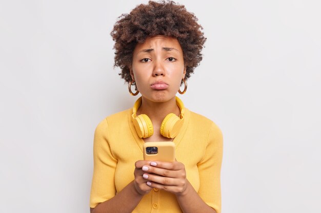 Mujer molesta frunce los labios mira tristemente al frente recibe un mensaje desagradable o una notificación en el teléfono móvil vestido de manera informal usa auriculares inalámbricos estéreo aislados en la pared blanca