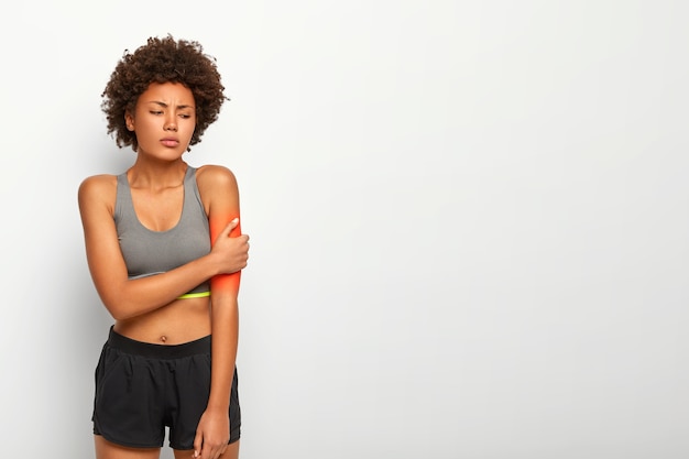 Mujer molesta toca el brazo, sufre de sentimientos dolorosos, mano herida durante el entrenamiento físico, vestida con blusa casual y pantalones cortos, posa en el interior sobre una pared blanca del estudio