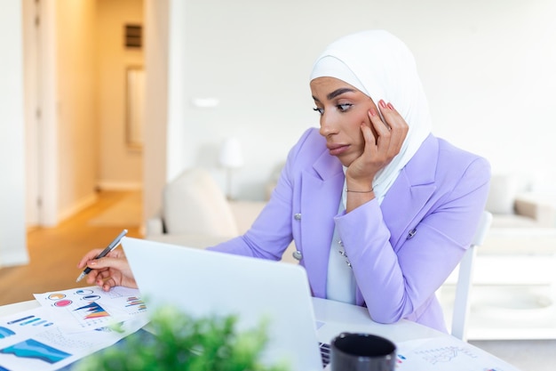 Mujer molesta usando hiyab frente a la búsqueda de computadoras portátiles y haciendo trabajo de oficina finanzas comerciales y concepto de estación de trabajo Empresaria árabe trabajando en casa