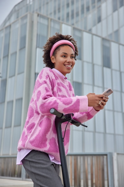 mujer monta scooter eléctrico por la ciudad tiene una expresión feliz vestida con ropa de moda usa transporte electrológico moderno y chats de teléfonos inteligentes en línea descubre nuevos lugares