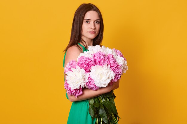 mujer morena abrazando gran ramo con peonías rosas y blancas, elegante mujer con flores, tiene expresión facial tranquila, posando aislado en amarillo.