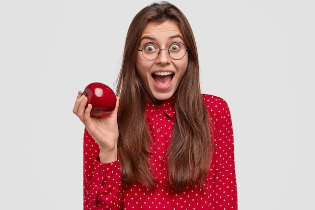 La mujer morena alegre abre la boca ampliamente, tiene una expresión feliz, usa gafas redondas, camisa de lunares rojos, sostiene una manzana fresca