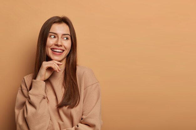 Una mujer morena alegre de aspecto agradable mira a un lado con una sonrisa encantadora, mantiene la mano debajo de la barbilla, usa una sudadera informal, posa contra la pared beige, recuerda un evento hilarante, tiene un aspecto alegre