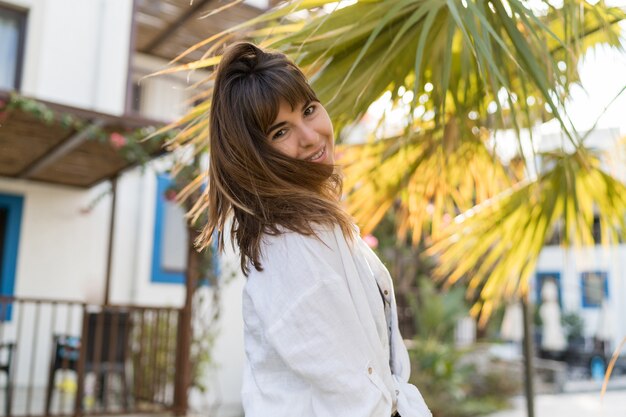 Mujer morena alegre disfrutando día de verano. Vistiendo blusa blanca. Palmeras en el fondo.