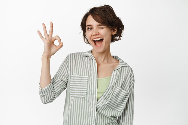 Mujer morena alegre guiñando un ojo y sonriendo, mostrando el signo OK OK, aprobar algo, elogiar y felicitar su elección, de pie sobre fondo blanco