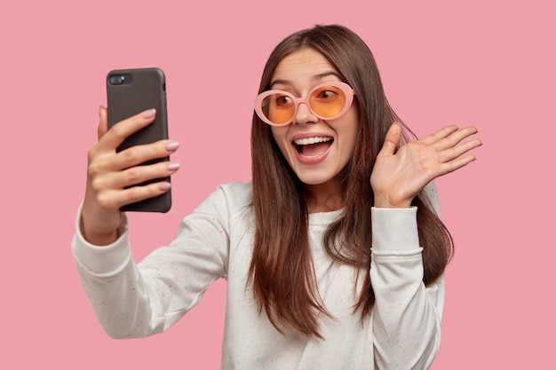 Mujer morena alegre lleva gafas de sol, hace videollamadas, conectado a internet inalámbrico, vestida con un jersey blanco, aislado sobre una pared rosa. Hola amigo