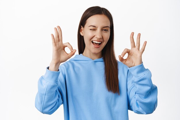 La mujer morena alegre muestra un gesto bien OK cero, guiña un ojo y sonríe satisfecho, dice que sí, alaba la gran elección, buen trabajo, excelente calidad, pared blanca.