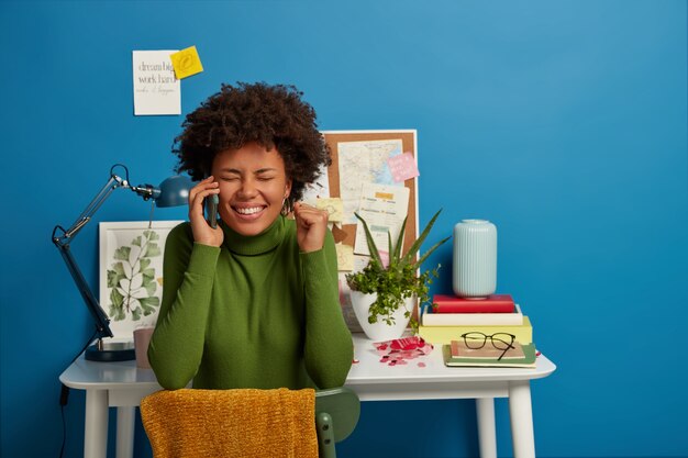 La mujer morena alegre tiene una conversación agradable, aplaude con el puño en alto, se regocija con el proyecto terminado, posa cerca del lugar de trabajo en casa, cierra los ojos de placer, aislado en la pared azul.