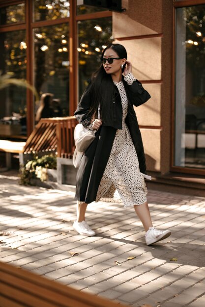 Mujer morena alegre con vestido de lunares midi y trinchera con estilo negro se mueve fuera Atractiva dama con gafas de sol camina por la calle