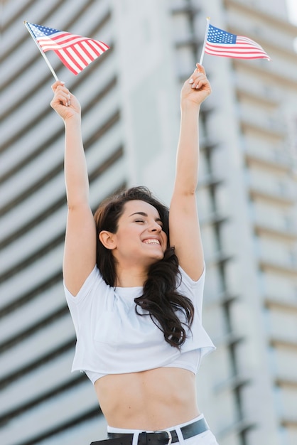 Foto gratuita mujer morena de ángulo bajo sosteniendo dos banderas de estados unidos y sonriendo