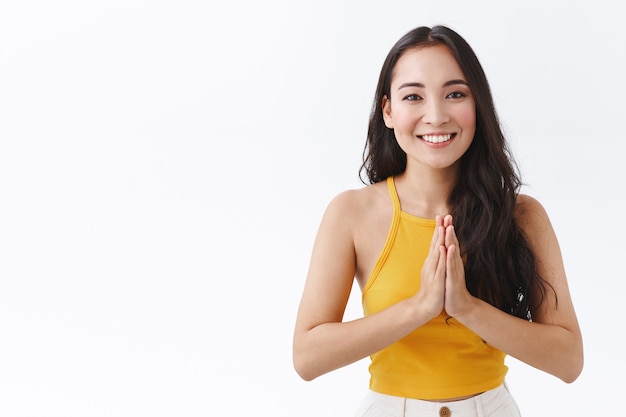 Mujer morena del este de Asia muy femenina en la parte superior amarilla de moda, juntar las manos en namaste, rogar o rezar gesto, sonriendo despreocupado, mirar con gratitud, agradeciendo por la ayuda, fondo blanco