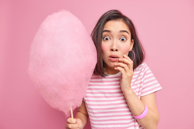 Mujer morena asustada aturdida sostiene hilo de caramelo dulce sorprendida al escuchar algo sorprendente y descubre cuántas calorías va a comer lleva una camiseta a rayas aislada sobre una pared rosa.