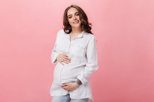 Mujer morena atractiva en camisa blanca y jeans sonríe y toca el vientre. Chica embarazada en jeans posa sobre fondo rosa.