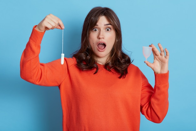 Mujer morena atractiva joven vistiendo ropa casual sosteniendo tampón y copa menstrual en las manos