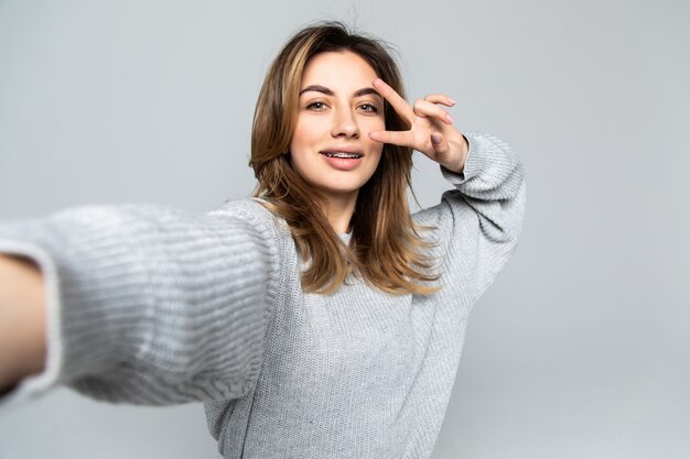 Mujer morena bastante alegre que muestra gesto de paz mientras toma selfie en teléfono inteligente, aislado sobre pared gris