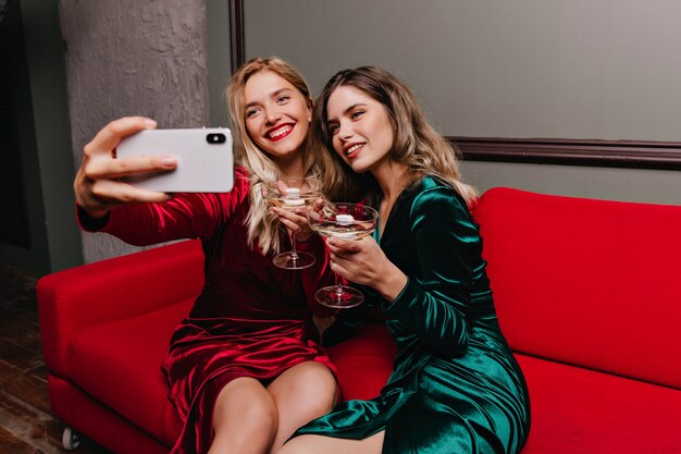 Mujer morena bebiendo vino mientras su amiga haciendo selfie. Retrato interior de chicas relajadas sentadas en el sofá rojo.