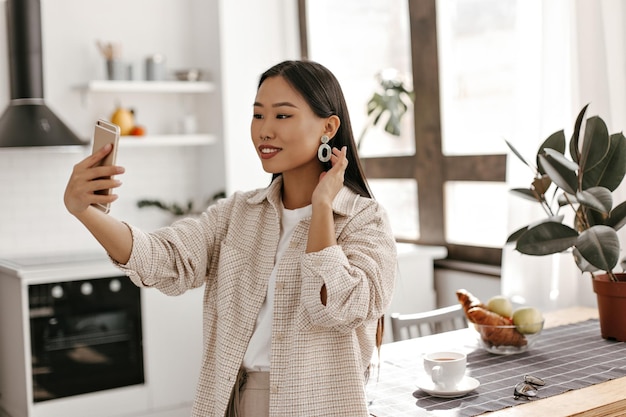 Mujer morena bronceada alegre con chaqueta beige se toma selfie en la cocina Dama asiática con traje elegante sostiene el teléfono y sonríe