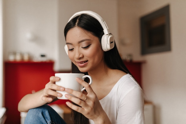 Mujer morena bronceada bebe té y escucha música con auriculares mientras está sentado en la cocina