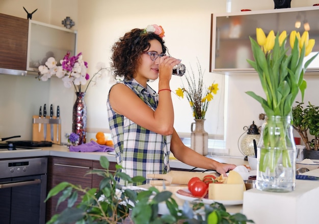 Mujer morena con cabello rizado bebe agua en una cocina.