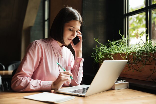 Mujer morena concentrada sentada junto a la mesa con la computadora portátil