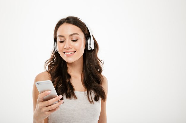 Mujer morena contenta en auriculares escuchando música y usando el teléfono inteligente sobre gris