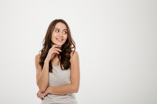 Mujer morena contenta posando con el brazo cerca de la cara y mirando a otro lado sobre gris