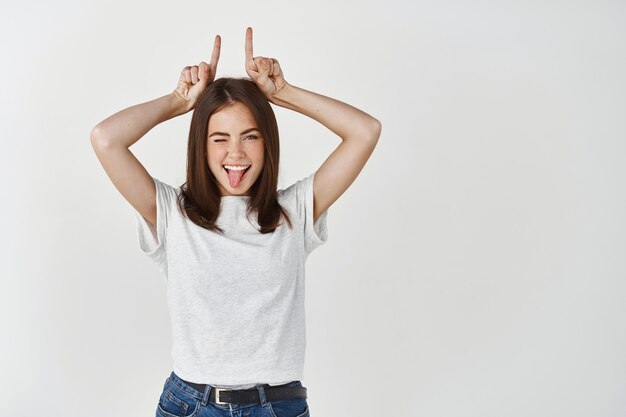 Mujer morena despreocupada mostrando cuernos de diablo con los dedos en la cabeza, sacando la lengua y sonriendo de alegría, de pie sobre una pared blanca.