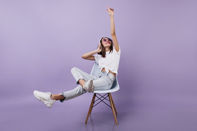 mujer morena divirtiéndose y escuchando música. Señorita bien vestida en auriculares sentado en una silla.