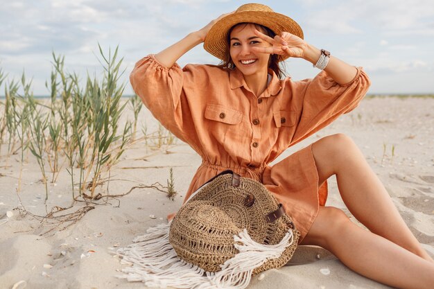 Mujer morena emocionada con sonrisa perfecta divirtiéndose en la playa soleada, sentada en la arena blanca cerca del océano.