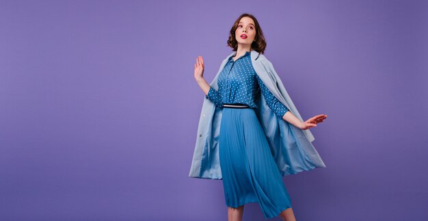 Mujer morena emocional en abrigo azul posando en la pared púrpura. Foto interior de hermosa modelo femenina de pelo corto en vestido midi de moda.