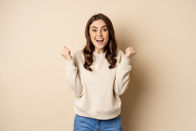 Mujer morena entusiasta riendo y sonriendo, animando, estrechando la mano y celebrando, triunfando, de pie sobre un fondo beige.