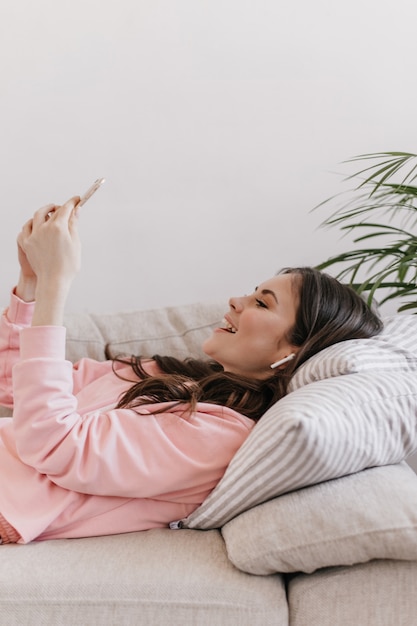 Mujer morena está acostada en el sofá y charlando con una sonrisa en el teléfono inteligente