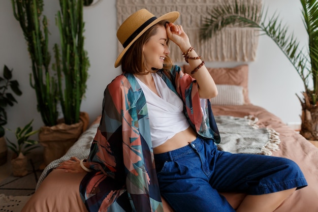 Mujer morena fascinante en sombrero de paja que se enfría en casa en el acogedor interior boho