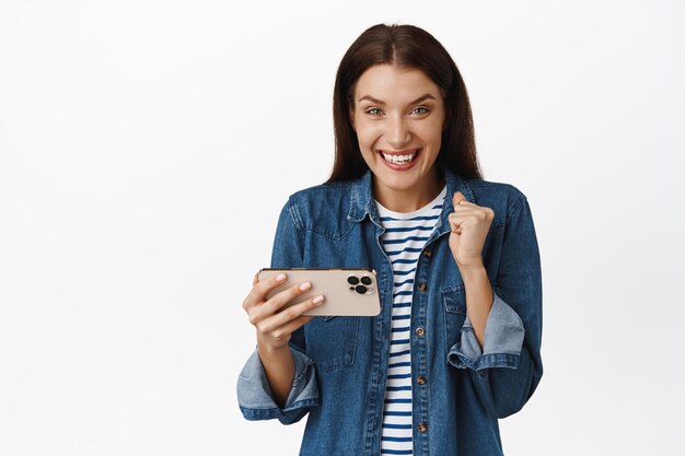 Mujer morena feliz ganando dinero por teléfono, jugando videojuegos móviles y regocijándose, celebrando el logro de objetivos en la aplicación, de pie contra el fondo blanco