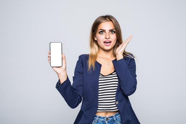 Mujer morena feliz sorprendida que muestra la pantalla del teléfono inteligente en blanco mientras mira con la boca abierta sobre la pared blanca