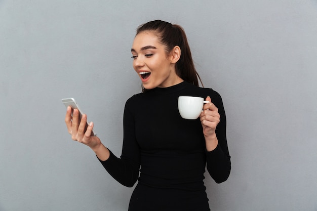 Mujer morena feliz sorprendida en ropa negra mirando smartphone