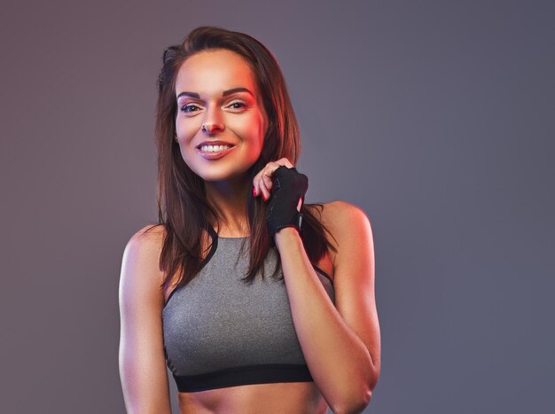 Mujer morena fitness sonriente en una ropa deportiva gris posando en un estudio. Aislado en un fondo gris.