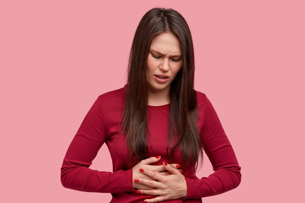 Mujer morena frustrada con expresión de disgusto, mantiene las manos en el vientre, siente dolor al igual que la menstruación, usa un suéter rojo