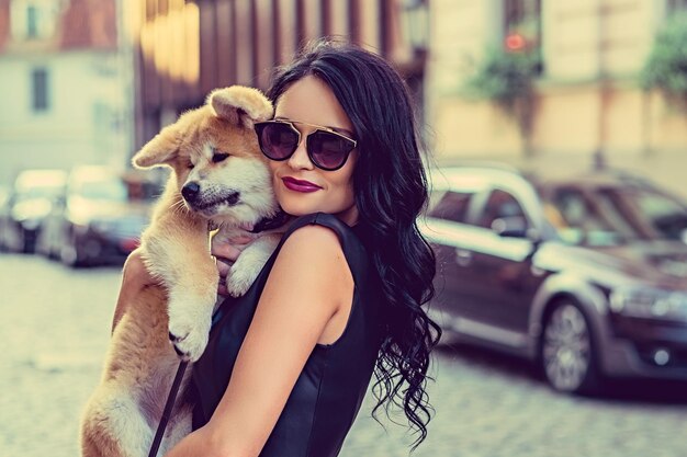 Mujer morena glamurosa con gafas de sol sosteniendo un cachorro de perro en una calle de un pueblo.