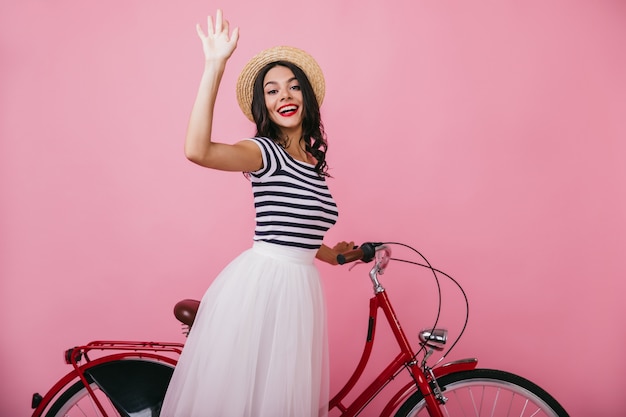 Mujer morena inspirada en camiseta sin mangas a rayas posando con bicicleta roja. Alegre dama bronceada de pie con una sonrisa.