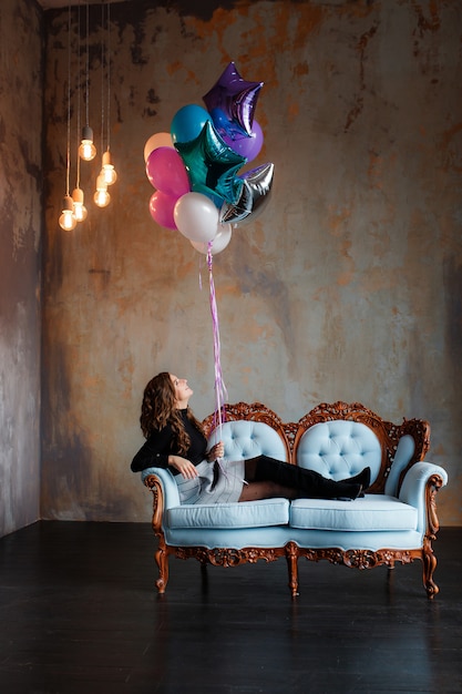 Mujer morena joven encantadora que sostiene un paquete grande de globos del helio que mienten en el sofá