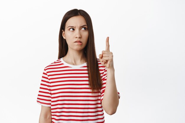 Mujer morena joven pensativa apuntando hacia arriba, mirando a un lado en el banner de venta y pensando, reflexionar sobre algo interesante, tener idea, de pie sobre la pared blanca.