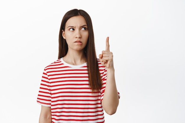 Mujer morena joven pensativa apuntando hacia arriba, mirando a un lado en el banner de venta y pensando, reflexionar sobre algo interesante, tener idea, de pie sobre la pared blanca.