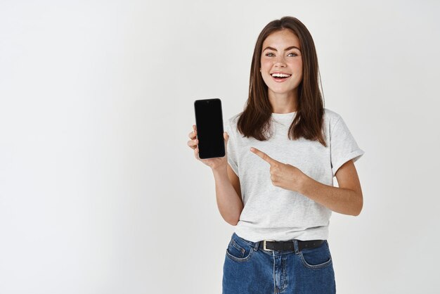Mujer morena joven que muestra la pantalla del teléfono inteligente en blanco y señala con el dedo el teléfono móvil sonriendo satisfecha con la cámara de pie sobre fondo blanco