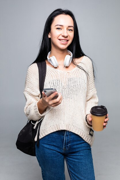 Mujer morena linda joven atractiva mientras usa el teléfono inteligente y bebe café aislado en la pared gris