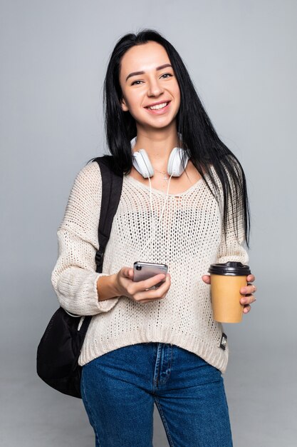 Mujer morena linda joven atractiva mientras usa el teléfono inteligente y bebe café aislado en la pared gris